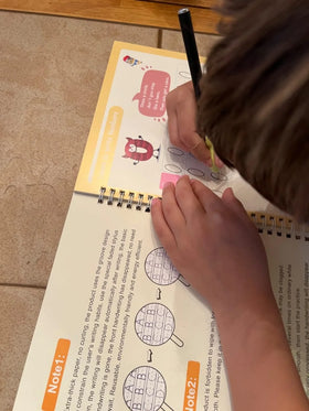 Review / child writing in a Tinker Mindz notebook with a pen on a wooden table.