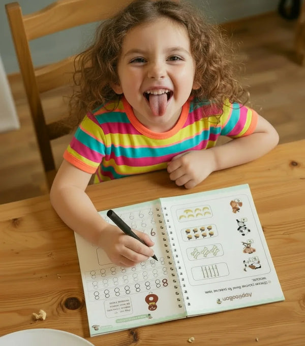 A happy young girl with curly hair wearing a rainbow striped shirt sits at a wooden table, playfully sticking her tongue out while tracing letters in a TinkerMindz reusable handwriting workbook.