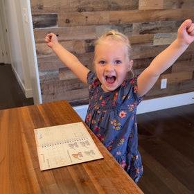 Child with arms raised in excitement near a wooden table with a notebook.
Customer review - 4-year-old girl's handwriting improvement using TinkerMindz grooved workbooks with before and after photos