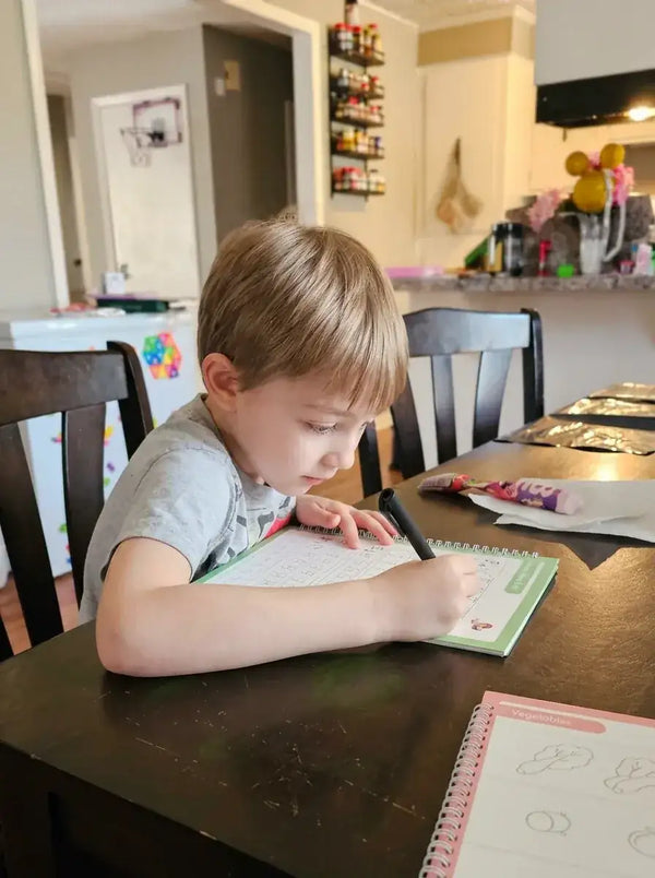 Child sitting at a table with a handwriting practice TinkerMindz copybook and crayons in a home setting.