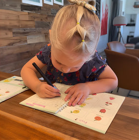 Child writing in a notebook with a pen on a wooden table.
Customer review - 4-year-old girl's handwriting improvement using TinkerMindz grooved workbooks with before and after photos