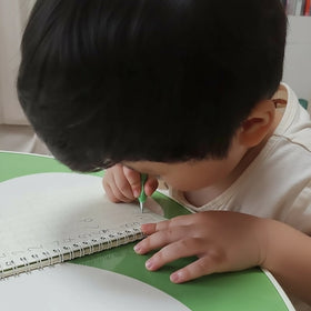 Review /  Child writing in a practice copybook with a pen and green grip on a green table.